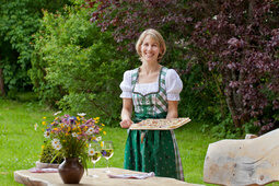 Eine lächelden Bedienung im grünen Kleid serviert Flammkuchen auf einem Holztablett im Garten des Hotel Konradshof.