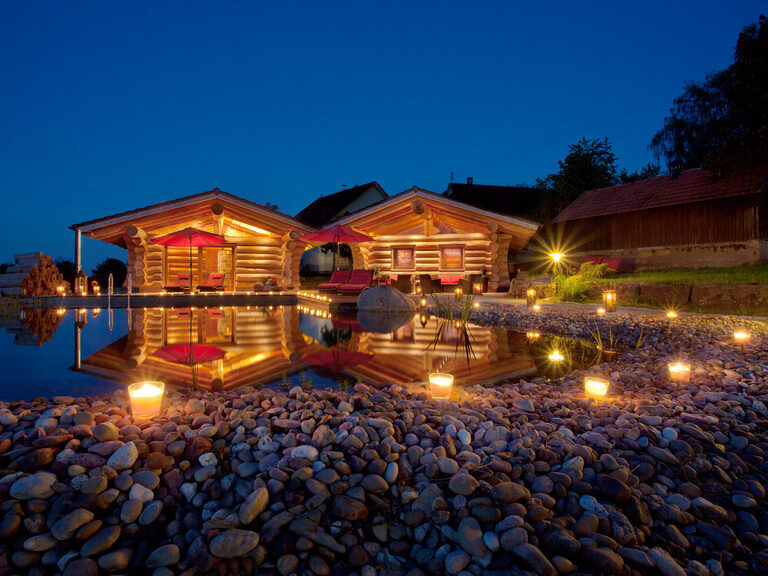 Zwei elegant beleuchtete Blockhütten an dem nächtlichen Natur-Schwimmteich in der Naturspa des Hotels Konradshof.